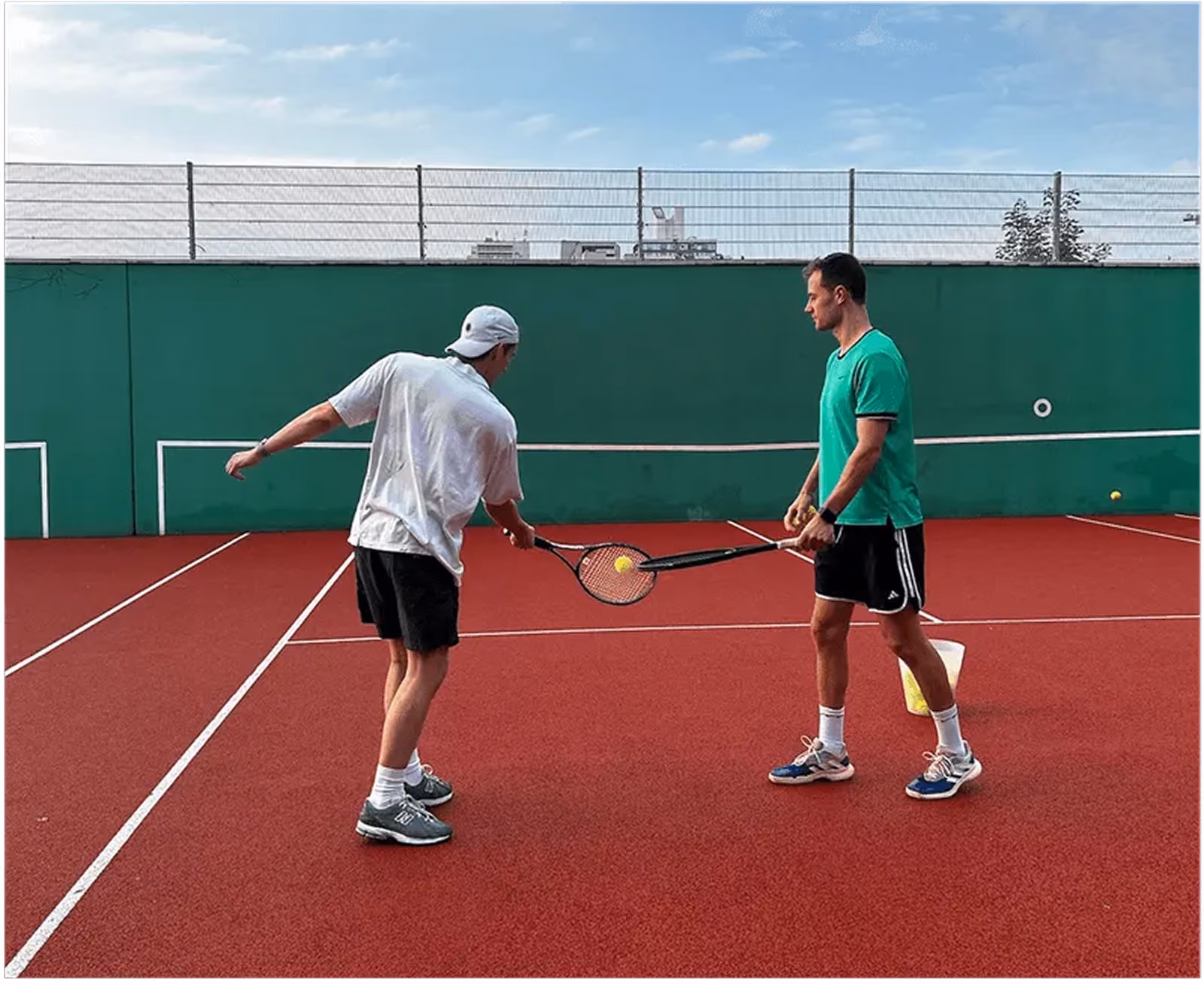 Zwei Männer auf einem Tennisplatz: Einer in weißem Shirt und Cap demonstriert mit einem Tennisschläger, der andere in grünem Shirt hält einen Schläger und Tennisbälle stehen in einem Eimer neben ihm.