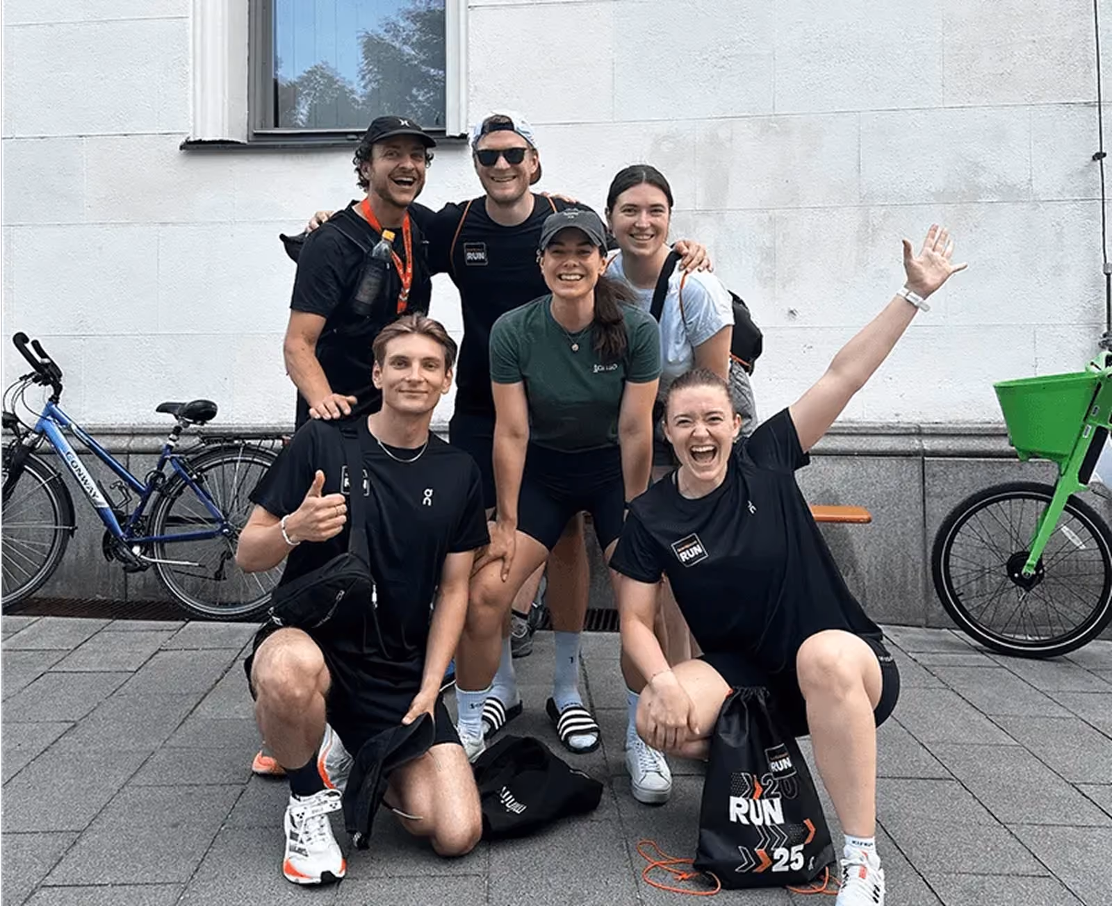 Eine Gruppe von sechs jungen Erwachsenen in sportlicher Kleidung posiert lachend vor einer weißen Wand, zwei von ihnen knien vorne, eine stellt den Daumen hoch, eine andere hebt die Hand jubelnd.