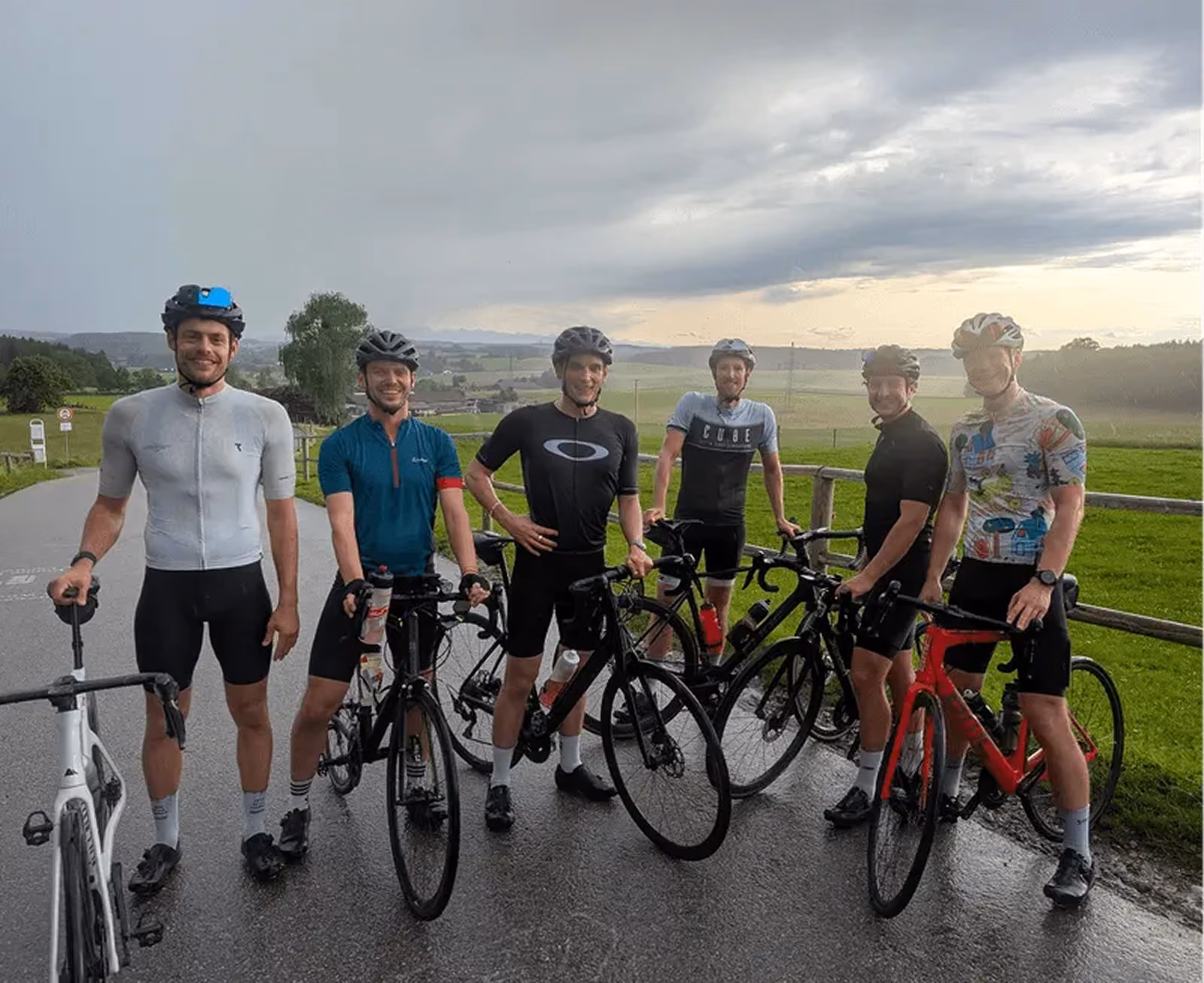 Sechs Radfahrer stehen mit ihren Rennrädern auf einer asphaltierten Landstraße vor einer ländlichen Kulisse mit grünen Wiesen und bewölktem Himmel.