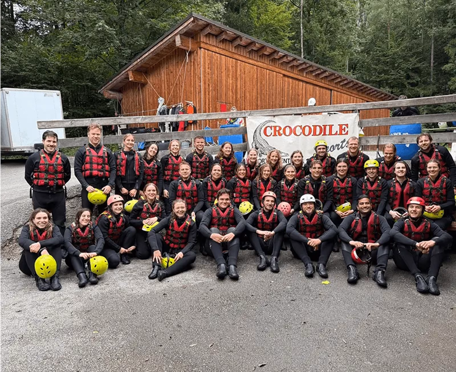Gruppenfoto von 35 Personen in Neoprenanzügen und Schwimmwesten vor einer Holzhütte mit einem Banner, das Crocodile Sports zeigt, einige halten Helme, umgeben von Wald.