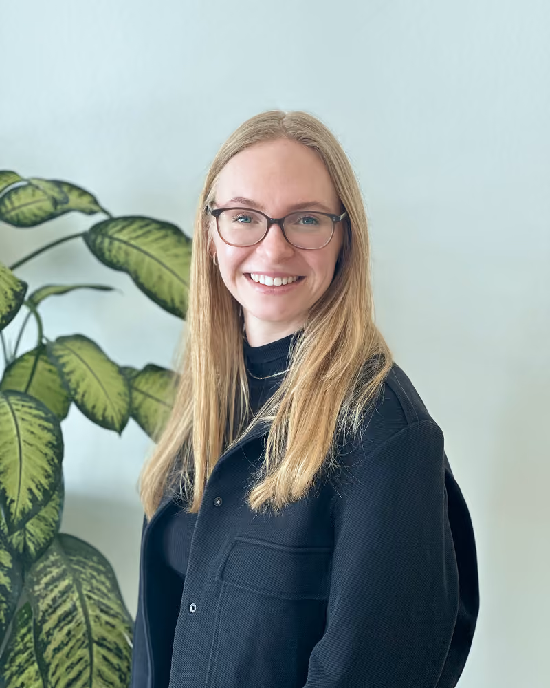 Frau mit langen blonden Haaren und Brille steht lächelnd vor einer hellen Wand, neben ihr eine große grüne Pflanze.