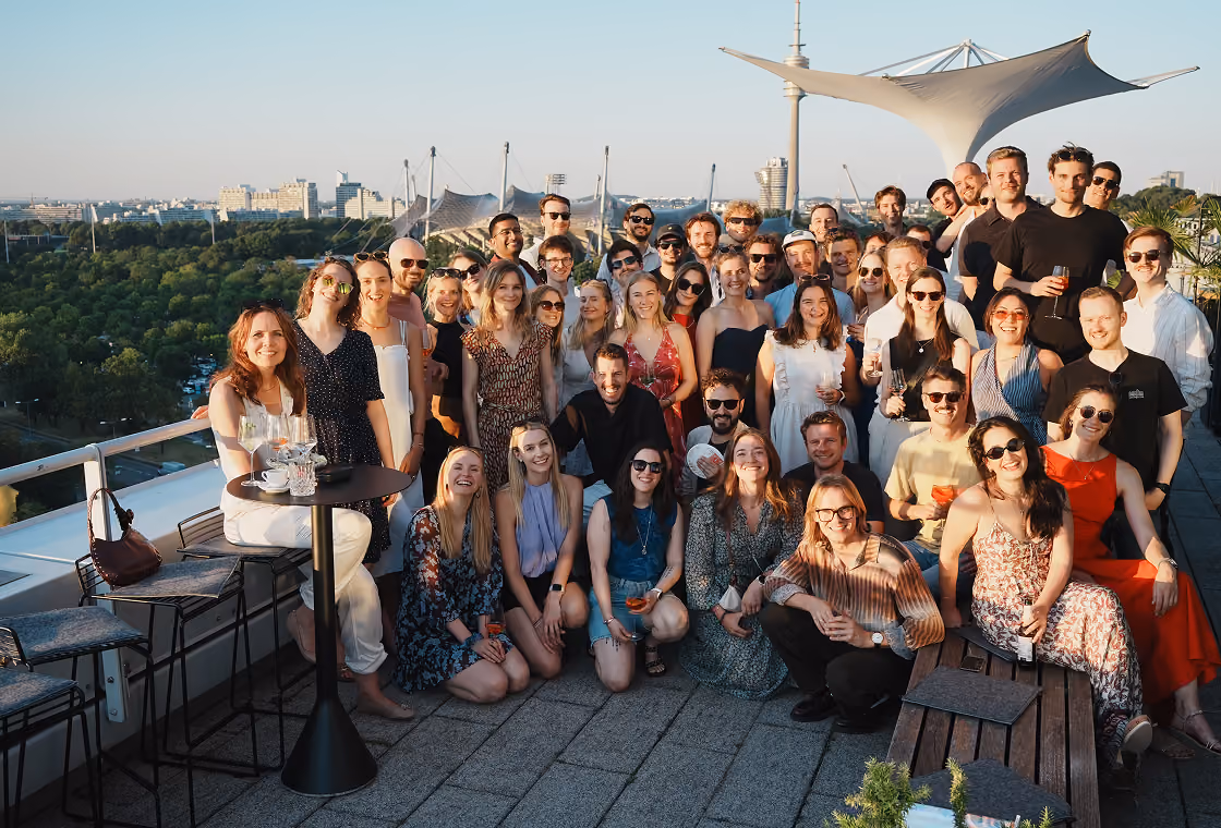Große Gruppe von Menschen lächelnd und posierend auf einer Dachterrasse mit Blick auf eine Stadt und grünen Park im Hintergrund bei Tageslicht, viele halten Getränke in der Hand.