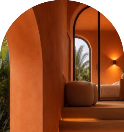 Interior view through an open archway showing a warm-toned room with terracotta-colored walls and ambient lighting under wooden steps leading up to the room with a round pouf and a bed, a large arched window with palm trees outside, and a wall sconce illuminating a vase on a bedside table.