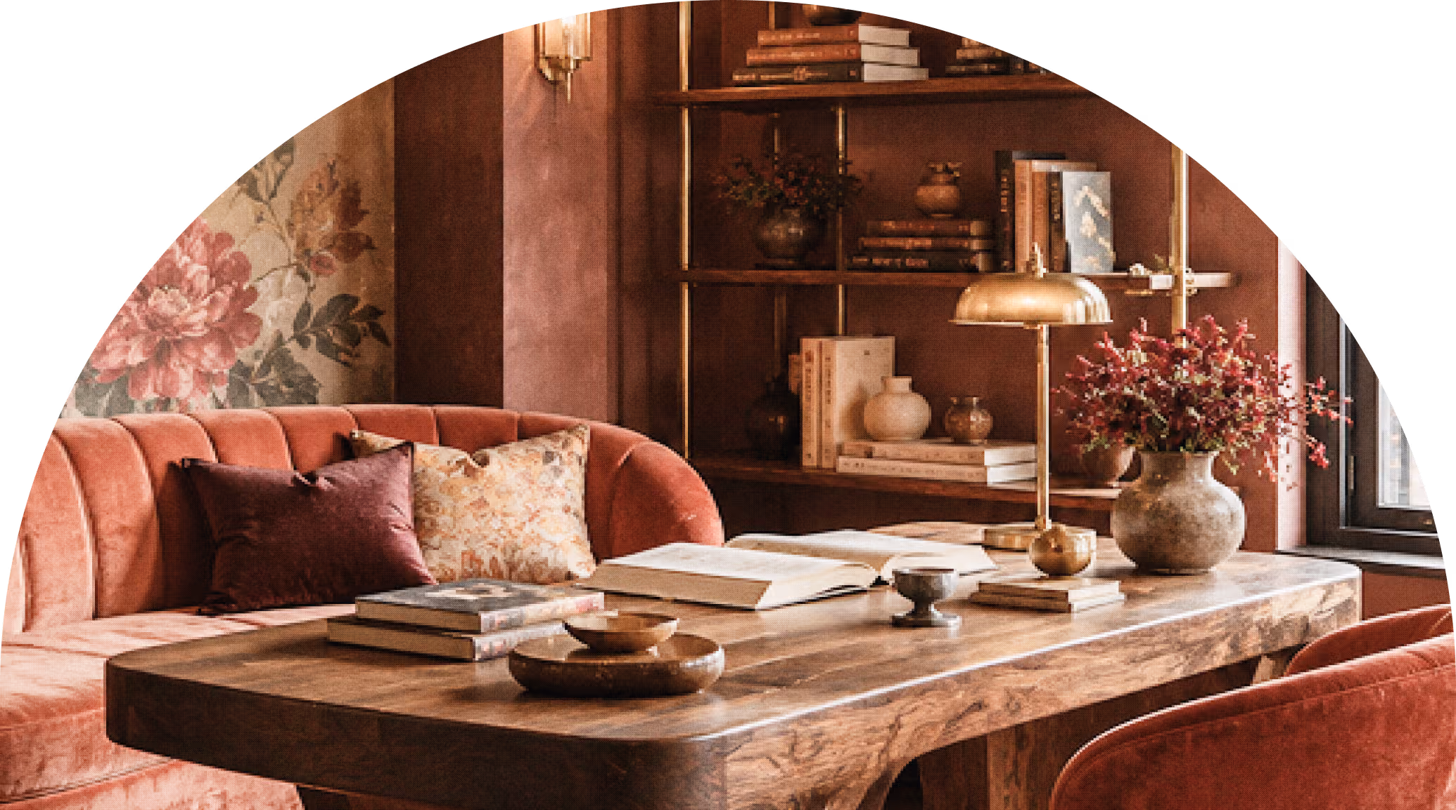 A cozy reading nook with a wooden table holding open books, a brass lamp, and a vase with red flowers, surrounded by a pink velvet tufted sofa and chair, with a floral curtain and a bookshelf filled with books and decorative items in the background.