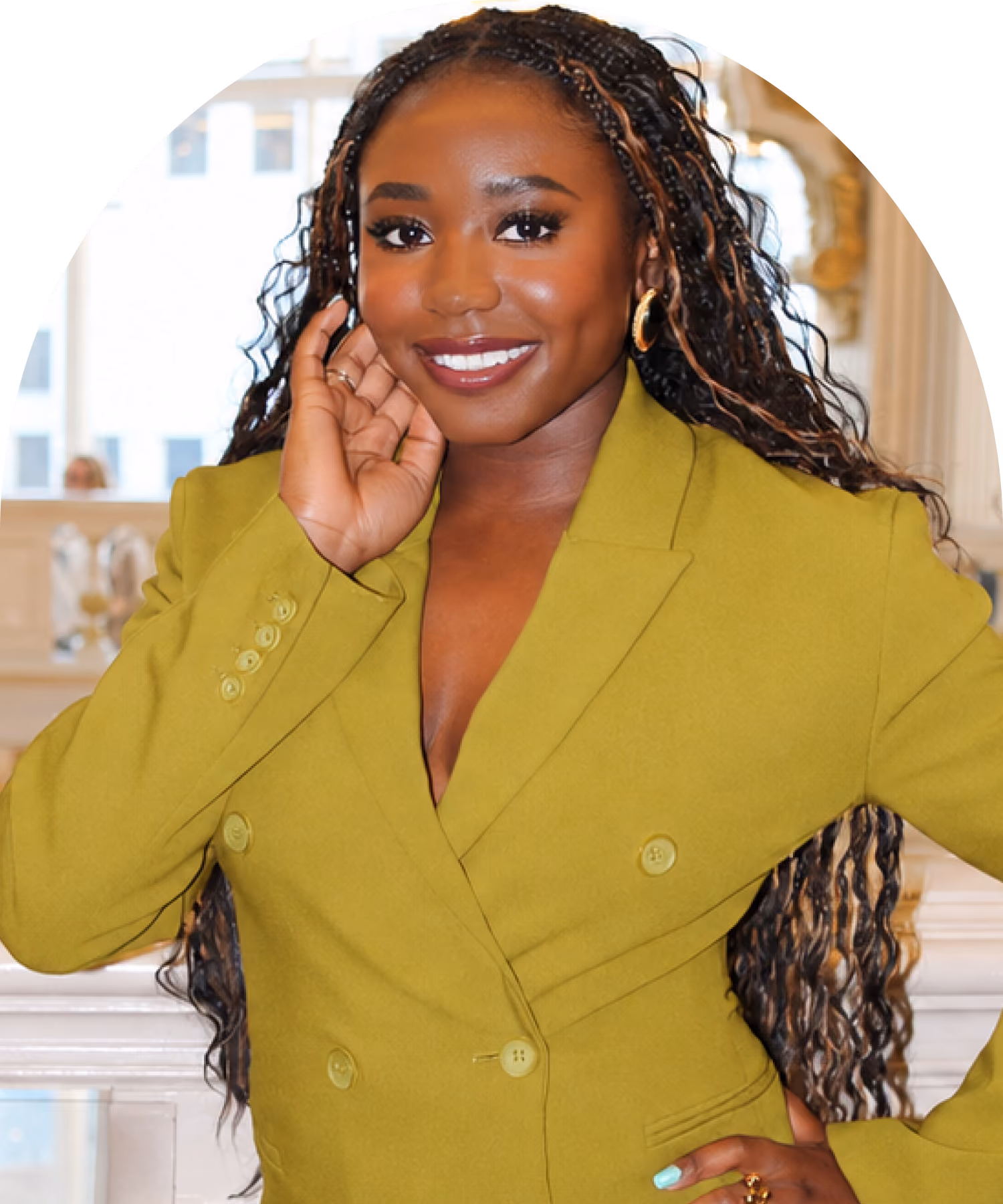 Smiling woman with long braided hair wearing a mustard yellow blazer, posing indoors with one hand touching her face and the other on her hip.