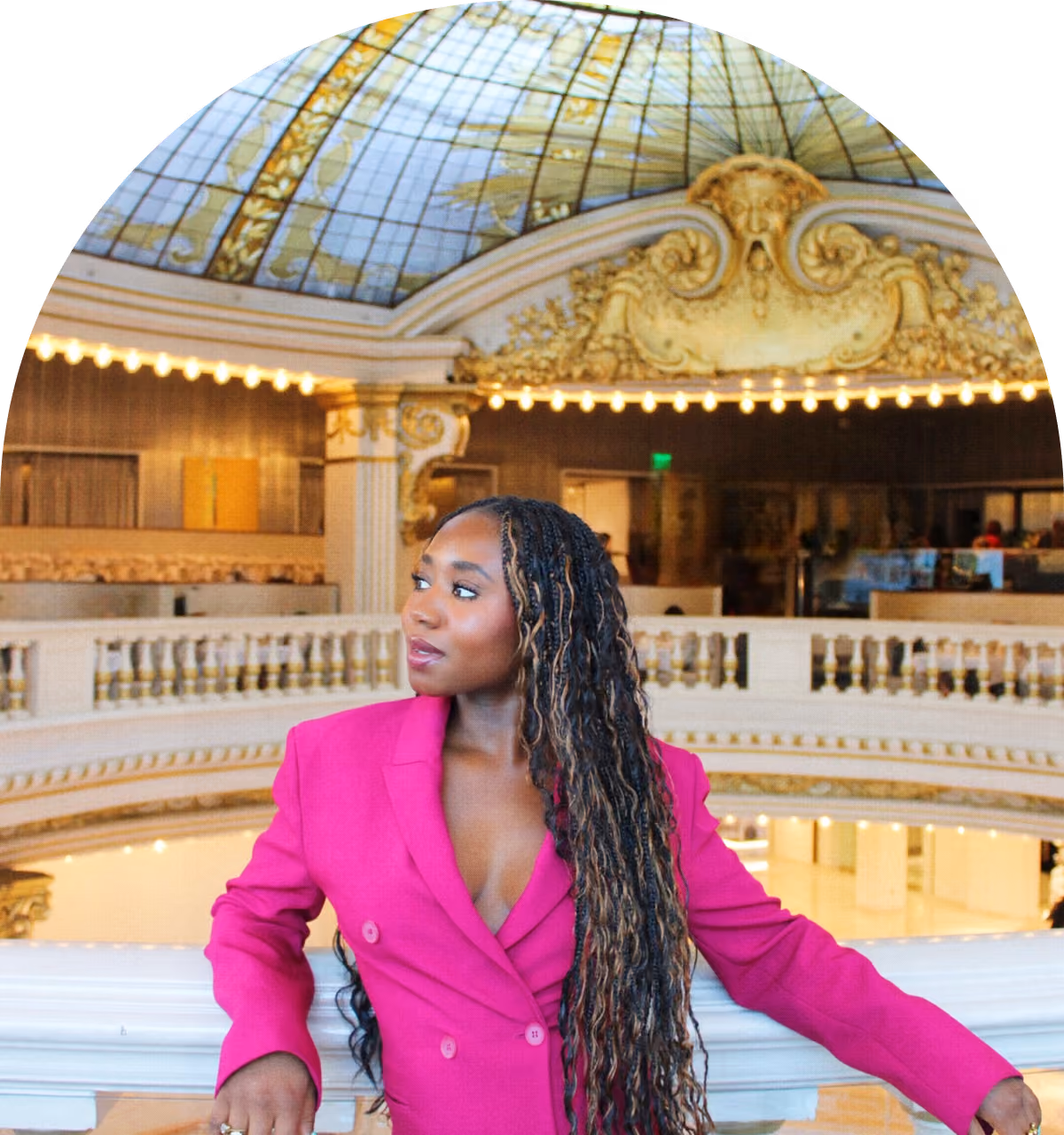 A woman with long braided hair and wearing a bright pink blazer stands indoors against a white railing, looking to her left, with an ornate ceiling and warm lighting in the background.