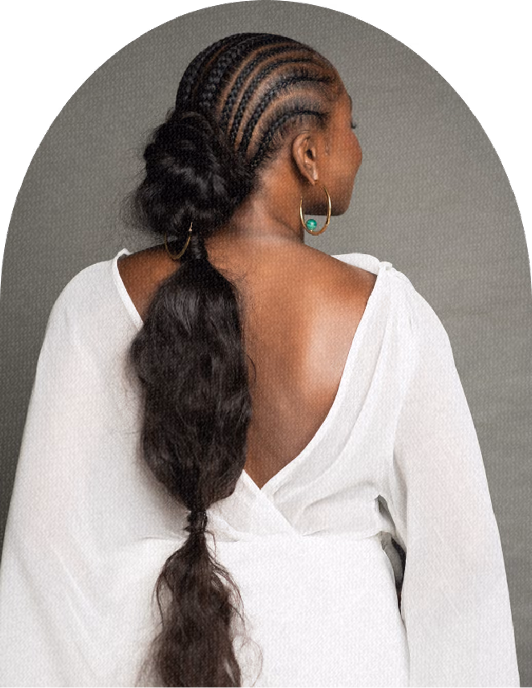 Woman with her back to the camera wearing a white top with an open back and styled hair in cornrow braids leading to a voluminous ponytail, adorned with hoop earrings.