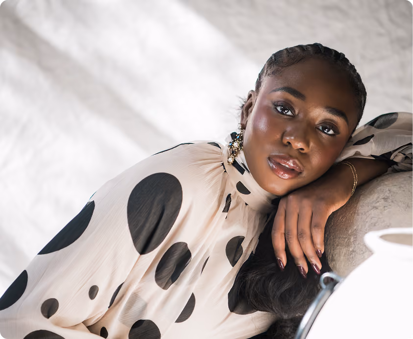 A woman with braided hair leans her head on her hand, wearing a cream blouse with large black polka dots and decorative earrings, posing against a light, textured background.
