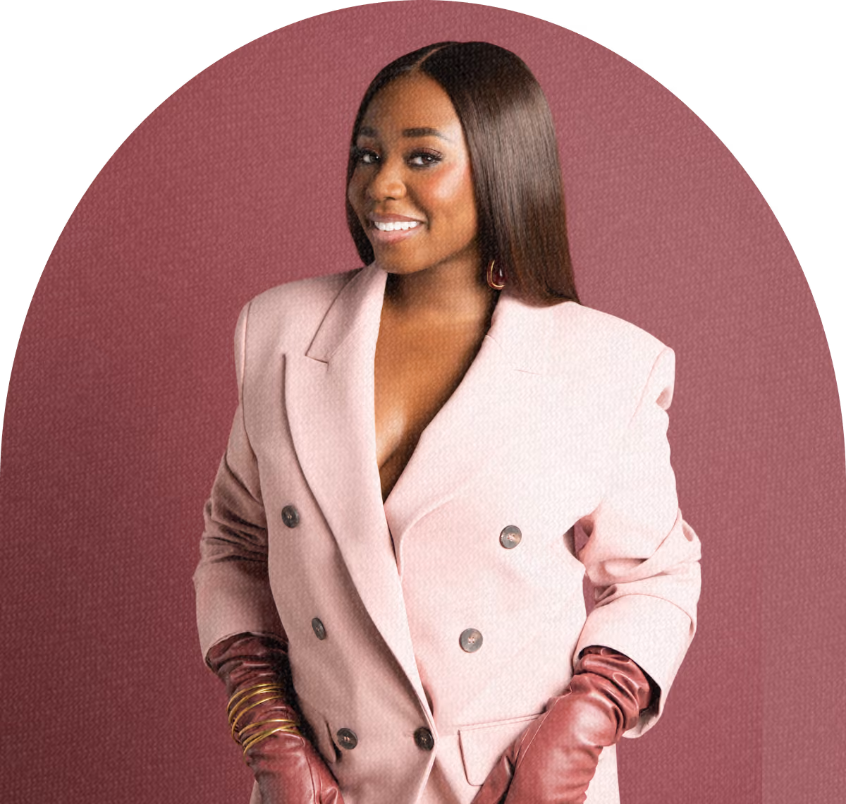 Smiling woman with long straight hair wearing a light pink double-breasted blazer and maroon leather gloves standing against a maroon background.