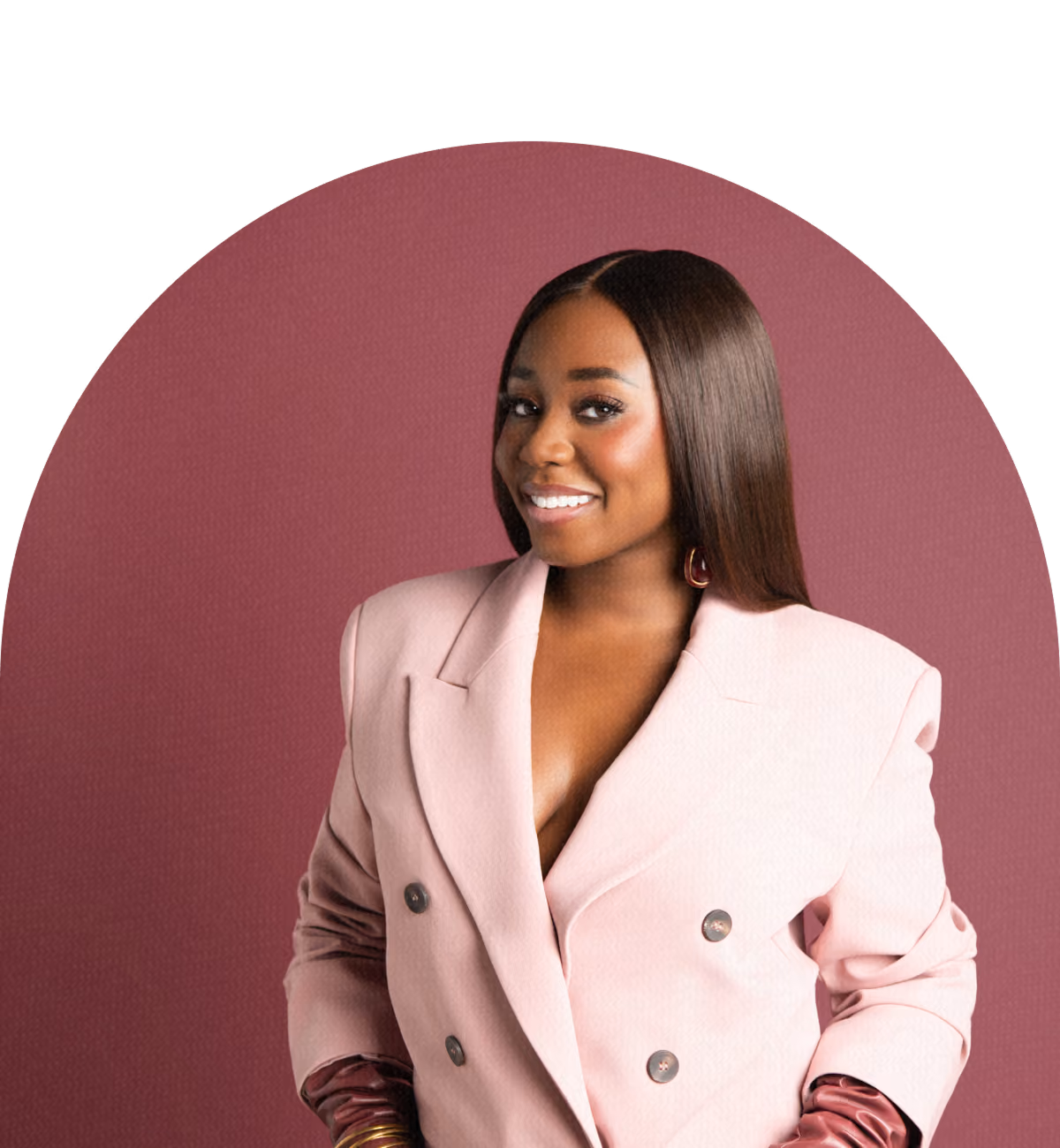 Smiling woman with long straight hair wearing a light pink double-breasted blazer with ruched sleeves standing against a mauve background.