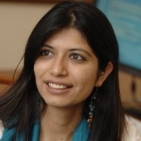 Smiling woman with long black hair, wearing dangling earrings and a teal scarf, indoors with blurred background.