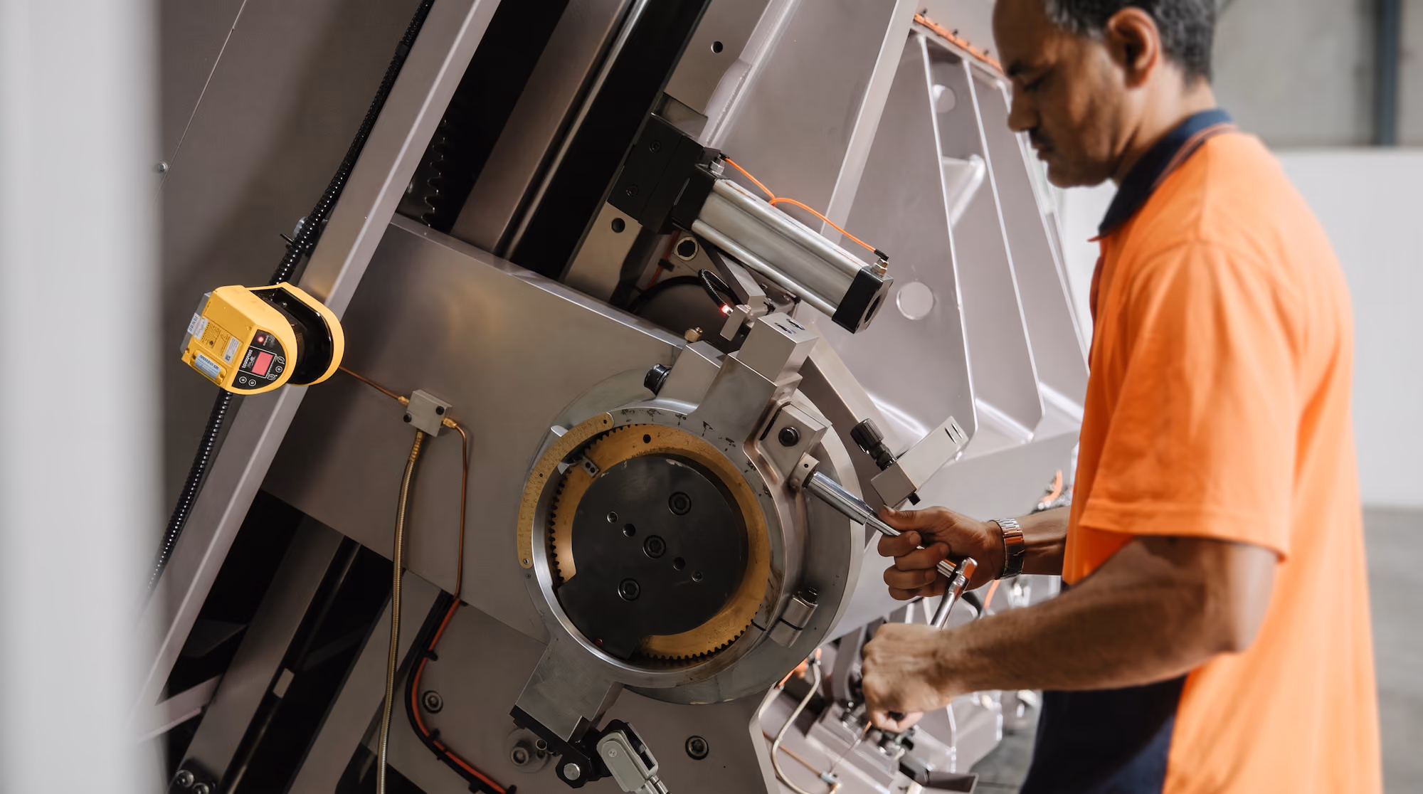 Man in an orange shirt operating or adjusting a large industrial machine with circular mechanical components and attached wiring.