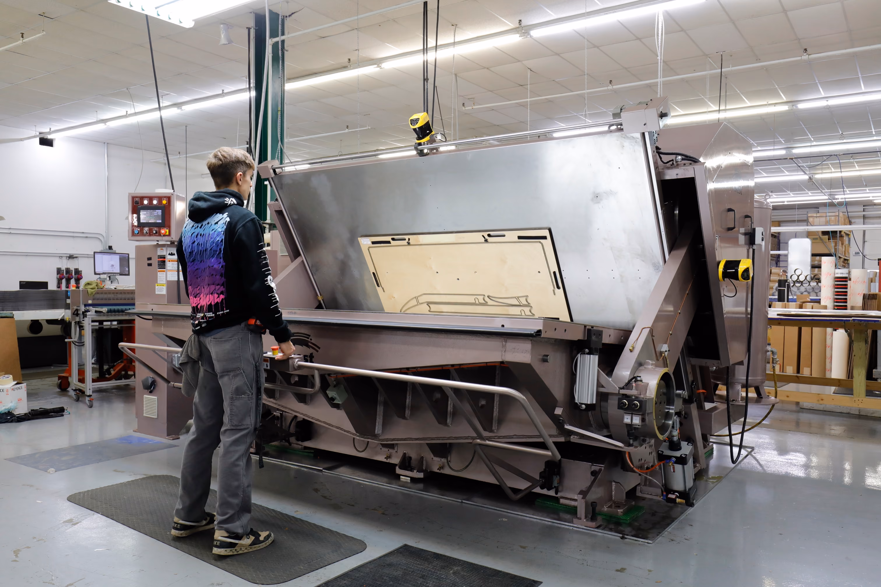 A person operating a large industrial machine in a factory setting with fluorescent overhead lighting and various equipment in the background.