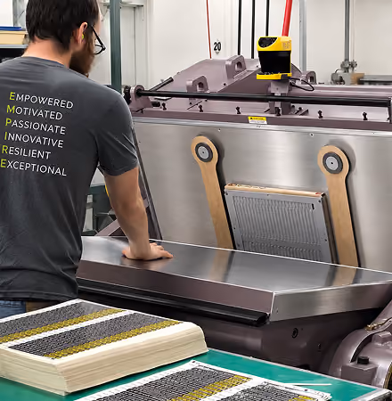 Man wearing a dark gray shirt with motivational words stands at a stainless steel industrial machine, examining printed sheets on a green table surface in a factory setting.