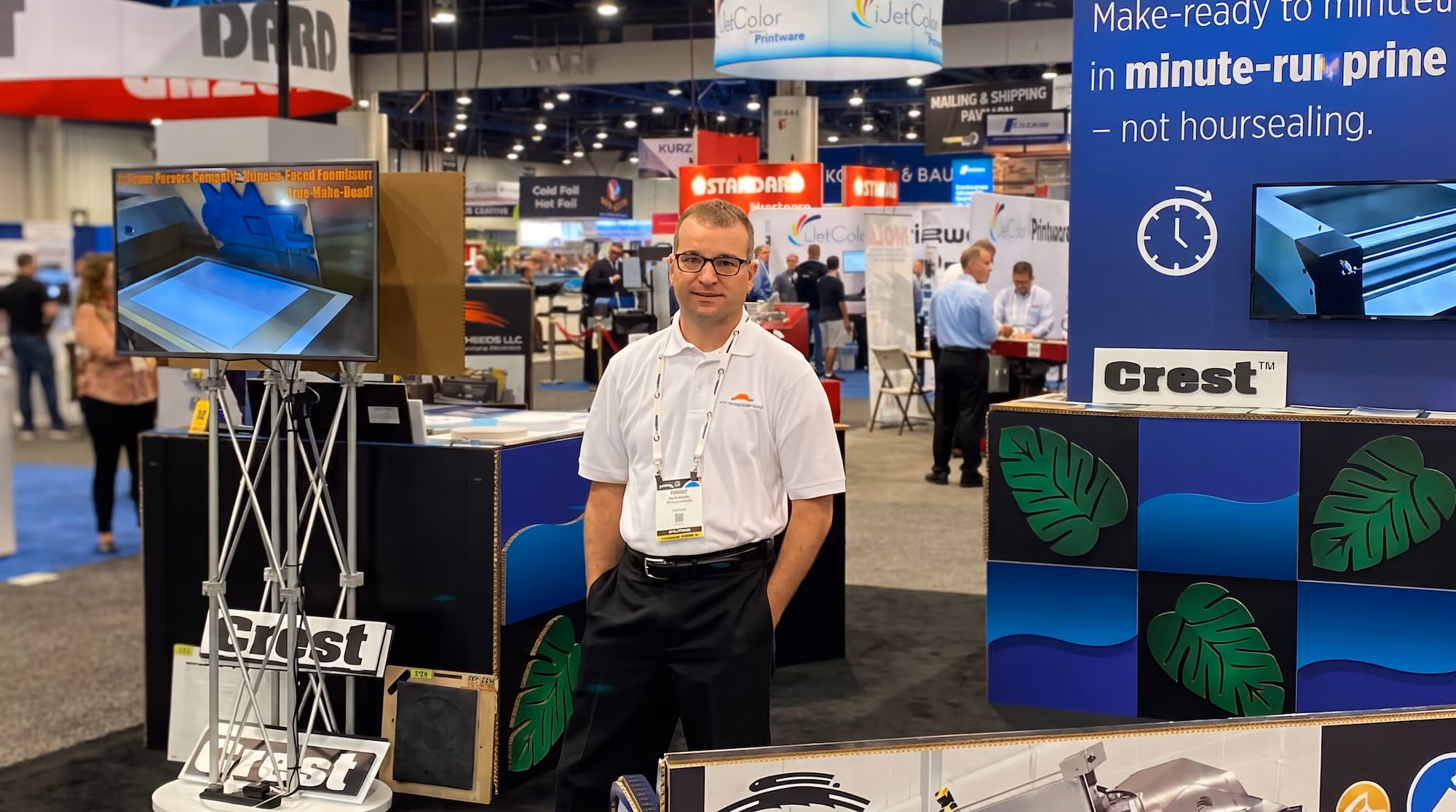 A man with glasses, wearing a white polo shirt and black pants, stands with hands in pockets at a convention booth displaying Crest branded promotional materials, with a monitor on a stand showing a machine interface.