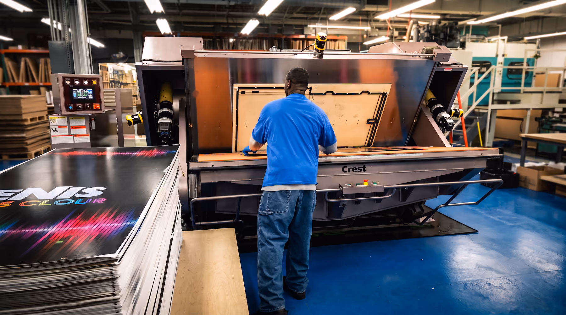 Man wearing a blue shirt and jeans operating a large printing machine labeled Crest in an industrial printing facility with stacks of printed sheets nearby.