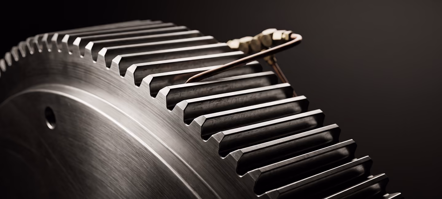Close-up of a large metallic gear with evenly spaced teeth and a small metallic wire or spring hooked around two teeth, set against a dark background.