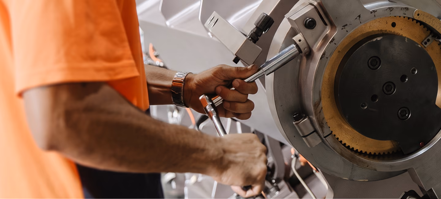 Person wearing an orange shirt adjusting or operating a piece of industrial machinery with visible gears and metal components.