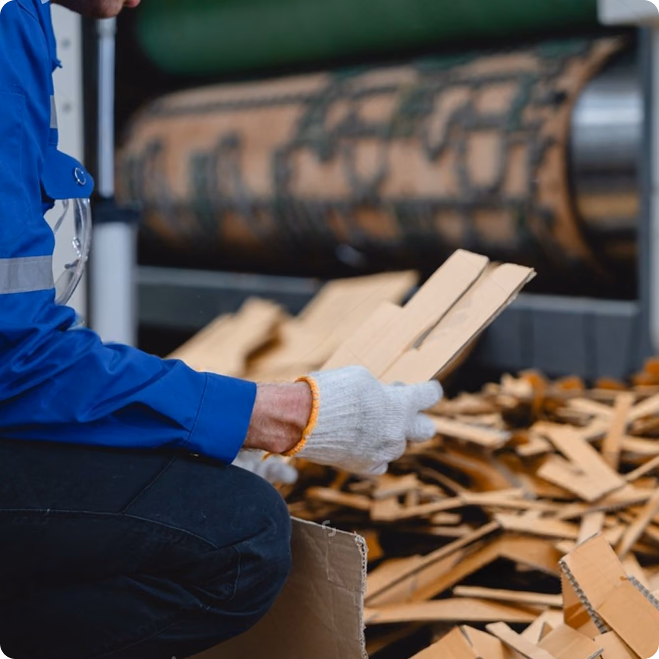 Person wearing a blue jacket and white gloves holding flat pieces of cardboard near a pile of cardboard scraps in an industrial setting.