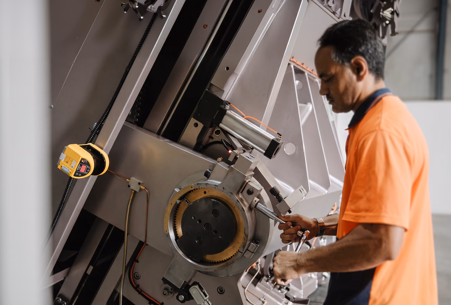 Man in an orange shirt operating or assembling a large industrial machine with mechanical and electronic components.