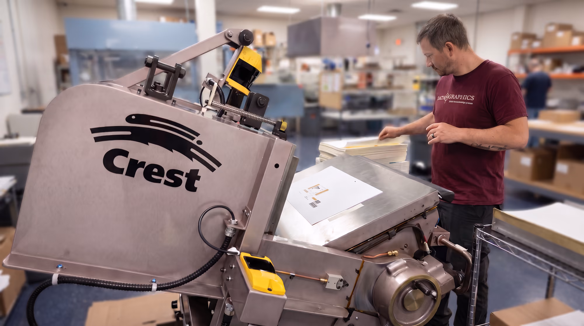 Man operating a large industrial machine labeled Crest in a busy workshop filled with shelves and boxes.