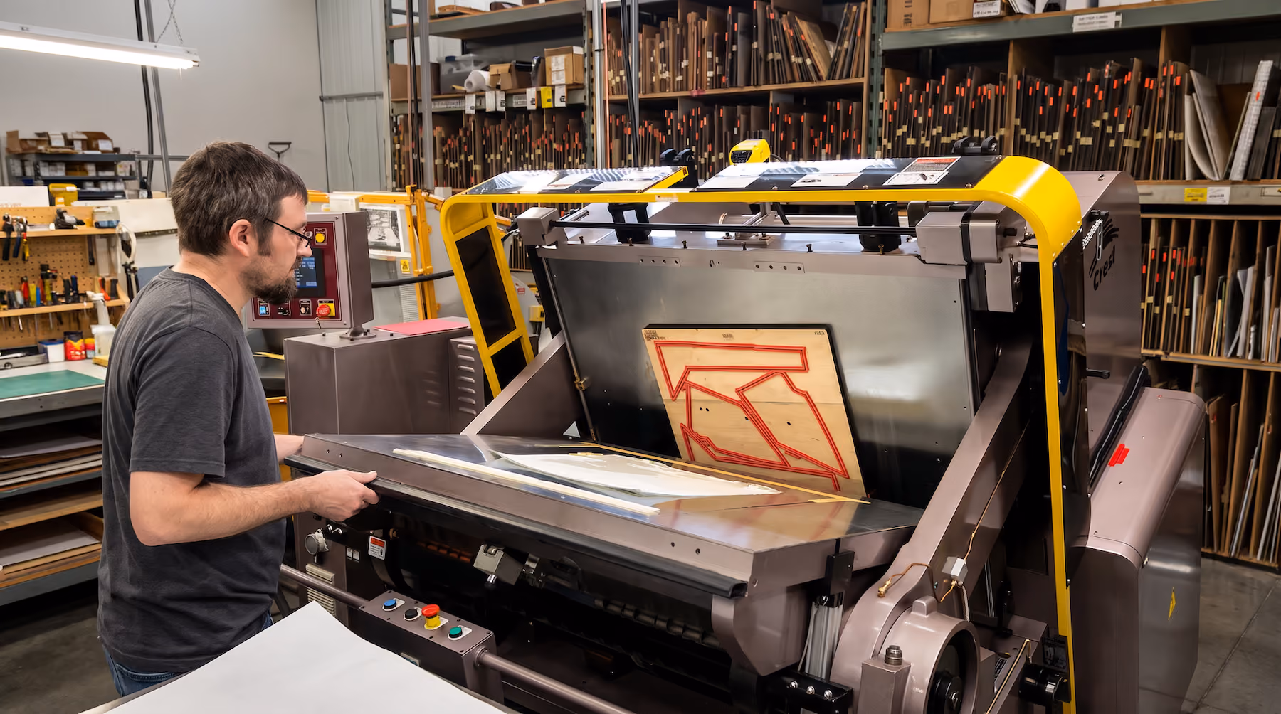 Man operating a large industrial machine in a workshop filled with shelves of rolled materials and tools.