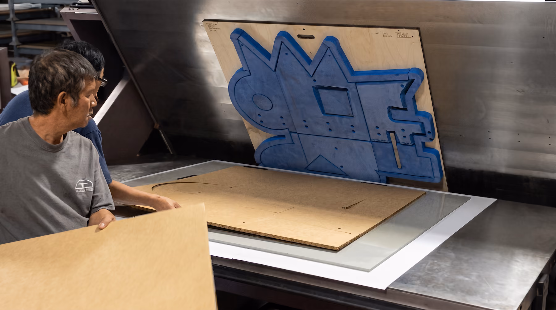 Two men working with large wooden stencil templates, one holding a sheet and the other near a machine with a metal surface and a vertical board featuring a complex geometric cutout design.