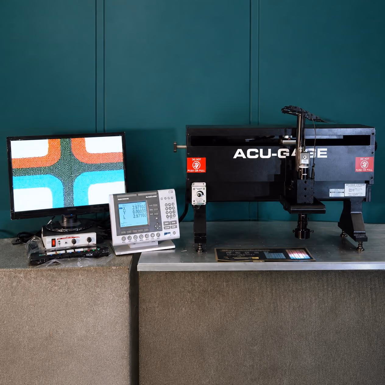 ACU-GAGE precision measuring equipment setup on a table including a black measurement device with red caution labels, a white control panel with numeric display, and a monitor showing a colorful patterned image.