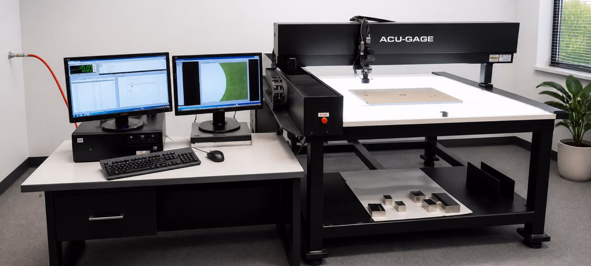 A large Acu-Gage inspection machine with a flat white surface and mechanical probe, positioned next to a desk holding two monitors displaying measurement software, a keyboard, and a mouse, in a bright room with a potted plant by the window.