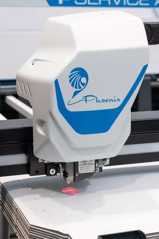Close-up of a white and blue automated cutting machine named Phoenix, with a red laser crosshair on stacked white sheets below it.