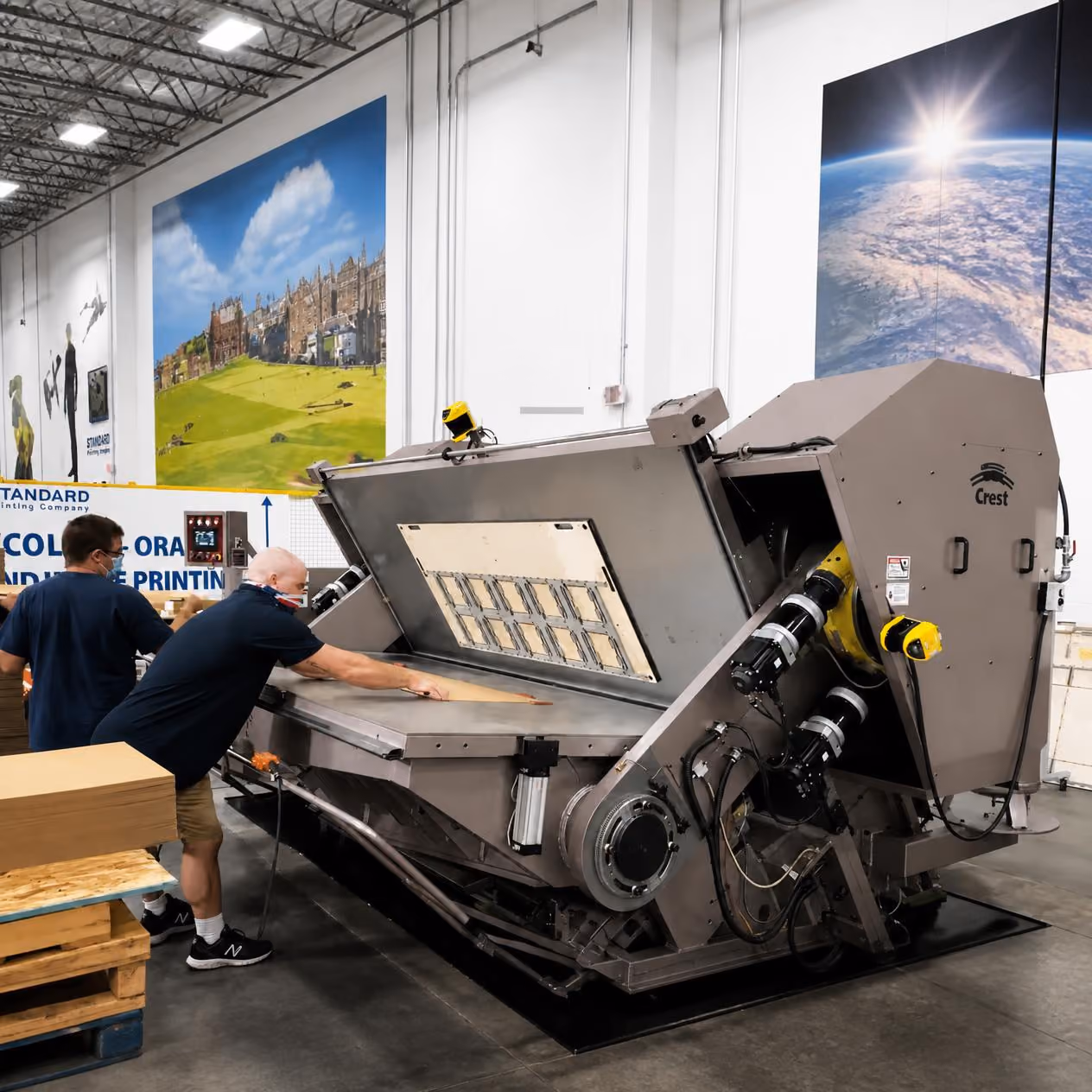Two men working with a large industrial printing or processing machine in a spacious warehouse with high ceilings and large wall art depicting landscapes and Earth.