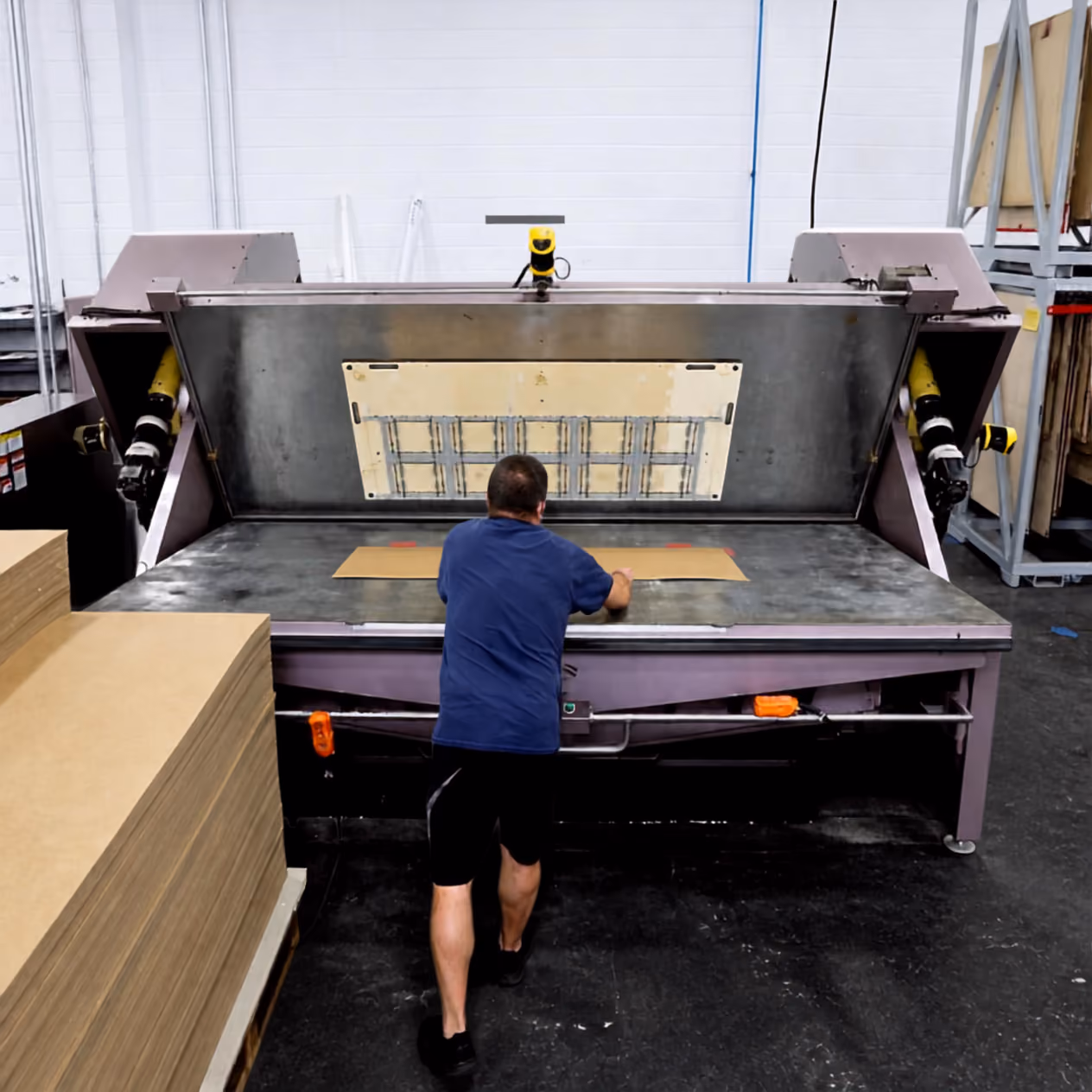 Man in a blue shirt and black shorts operating a large industrial machine with a flat surface and a raised lid in a warehouse setting.
