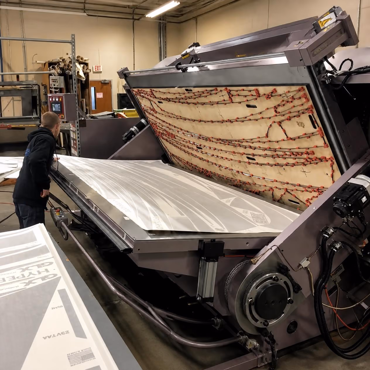 A man inspects a large industrial machine in a factory that is processing a sheet of material with printed text and lines, the machine has a tilted upper section with red wiring and mechanical components.