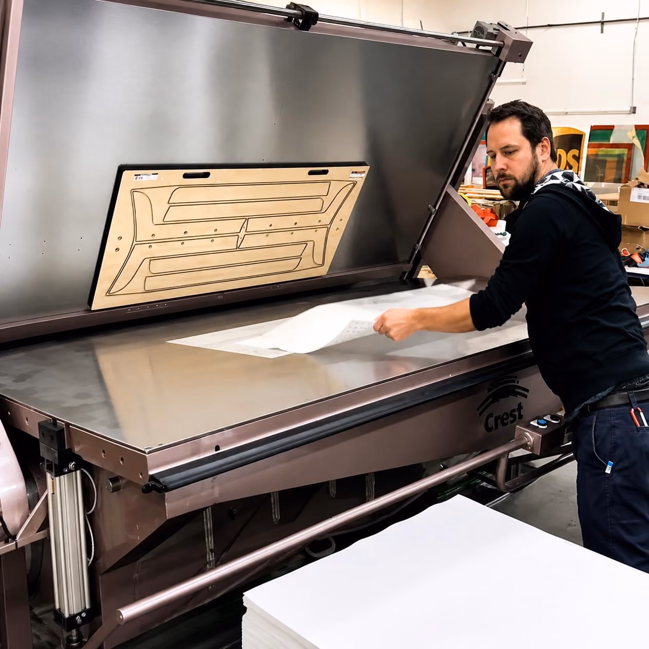 A man operating a large flatbed machine labeled 'Crest,' lifting a sheet of material off the machine's flat surface in an industrial workspace.