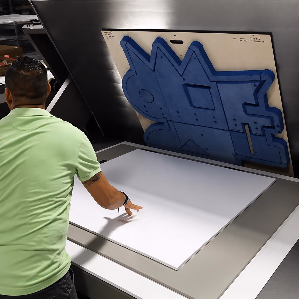 Man in a light green shirt operating a large industrial cutting machine with a blue foam die on the vertical panel and a large white sheet on the horizontal surface ready to be cut.