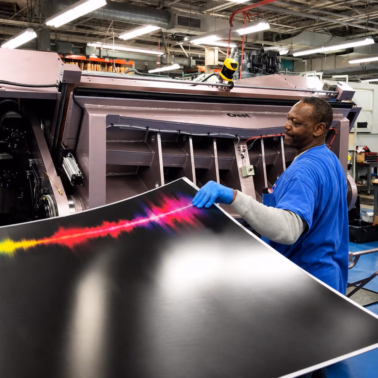 A man wearing blue gloves and a blue shirt inspects a large printed sheet with colorful waveforms in a factory setting beside a large industrial printing machine.