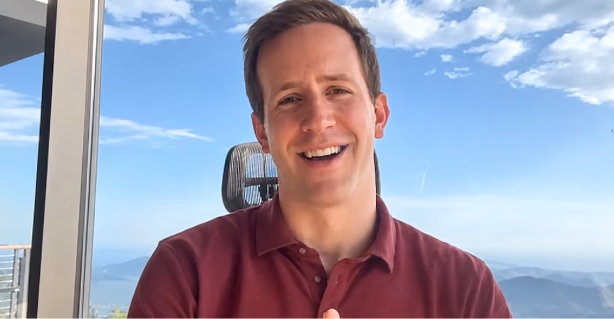 Smiling man in a maroon polo shirt sitting indoors near a window with a scenic mountain and blue sky background.
