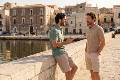 Two men casually dressed in summer clothes stand and lean against a stone railing by a waterfront, smiling and talking with historic buildings in the background.