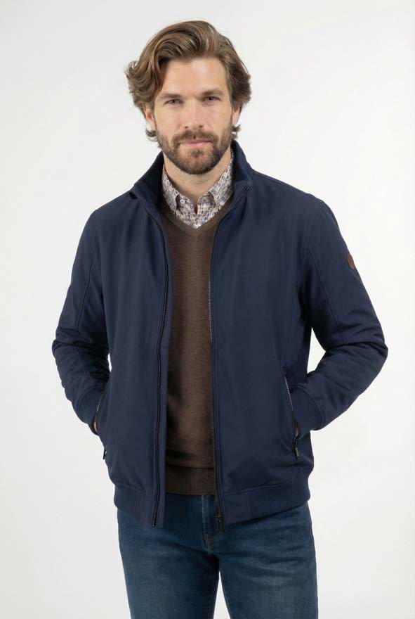 Bearded man with wavy brown hair wearing a navy blue jacket over a brown sweater and checkered shirt, standing with hands in pockets against a plain white background.