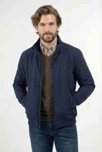 Man with light brown hair and beard wearing a navy jacket over a brown sweater and blue jeans, posing with hands in jacket pockets against a light background.