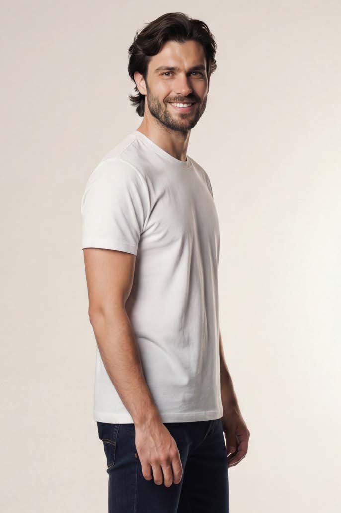 Smiling man with dark hair and beard wearing a plain white t-shirt and dark jeans standing sideways against a light beige background.