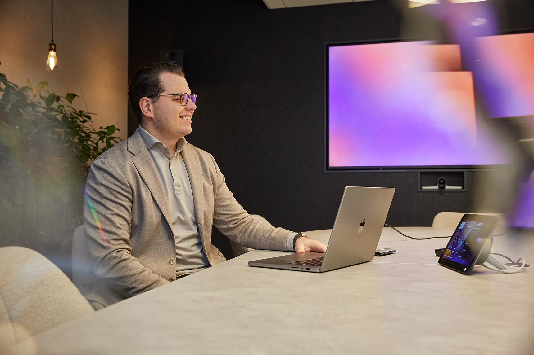 Man met bril in een beige colbert aan een vergadertafel met laptop en tablet, met op de achtergrond een scherm met een paars-oranje verloop