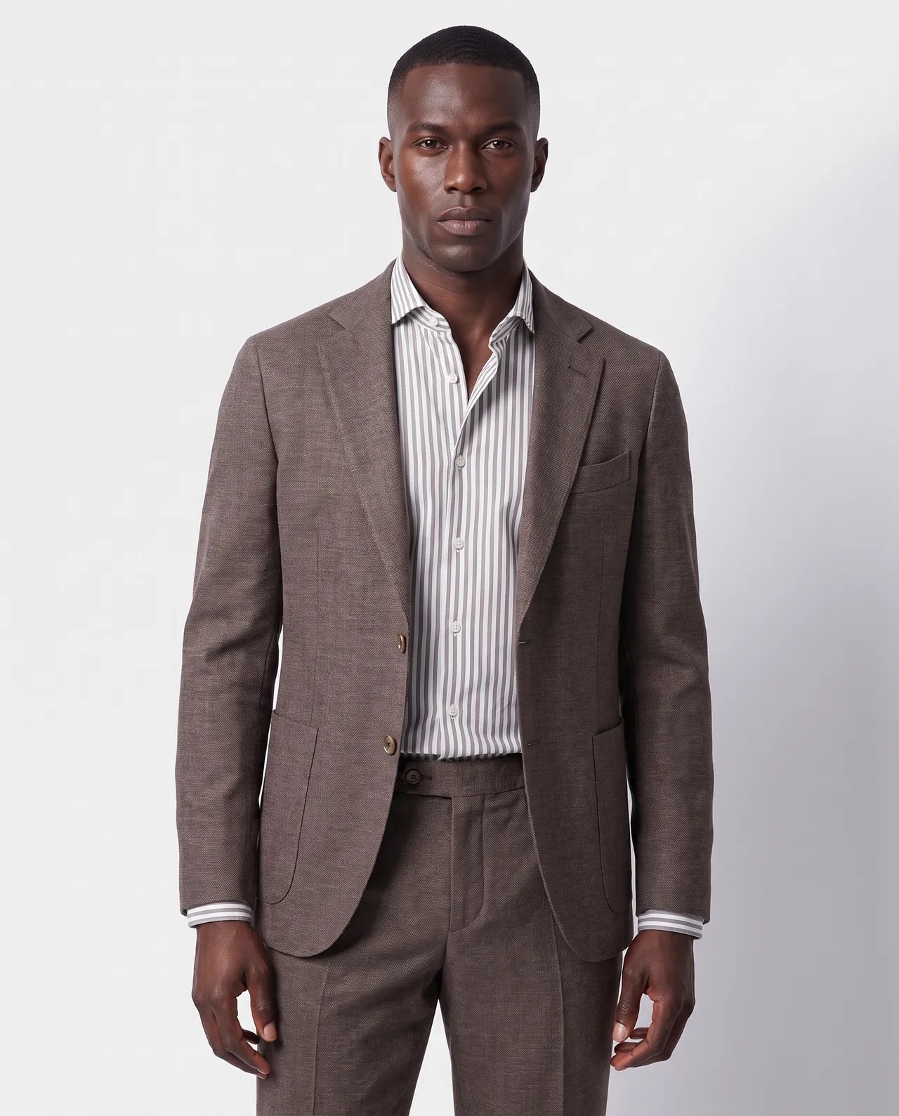 Man wearing a brown suit jacket and matching trousers over a white dress shirt with thin vertical gray stripes, standing against a plain light gray background.