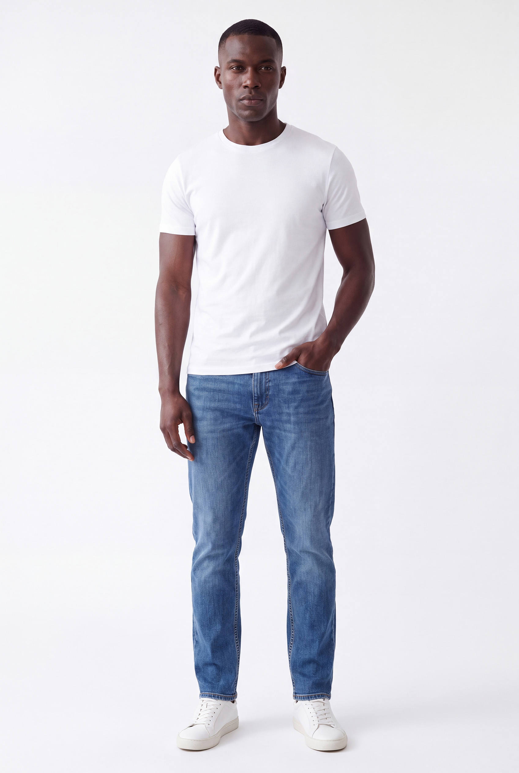 Full-length photo of a man standing against a white background wearing a plain white t-shirt, blue jeans, and white sneakers with one hand in his pocket.