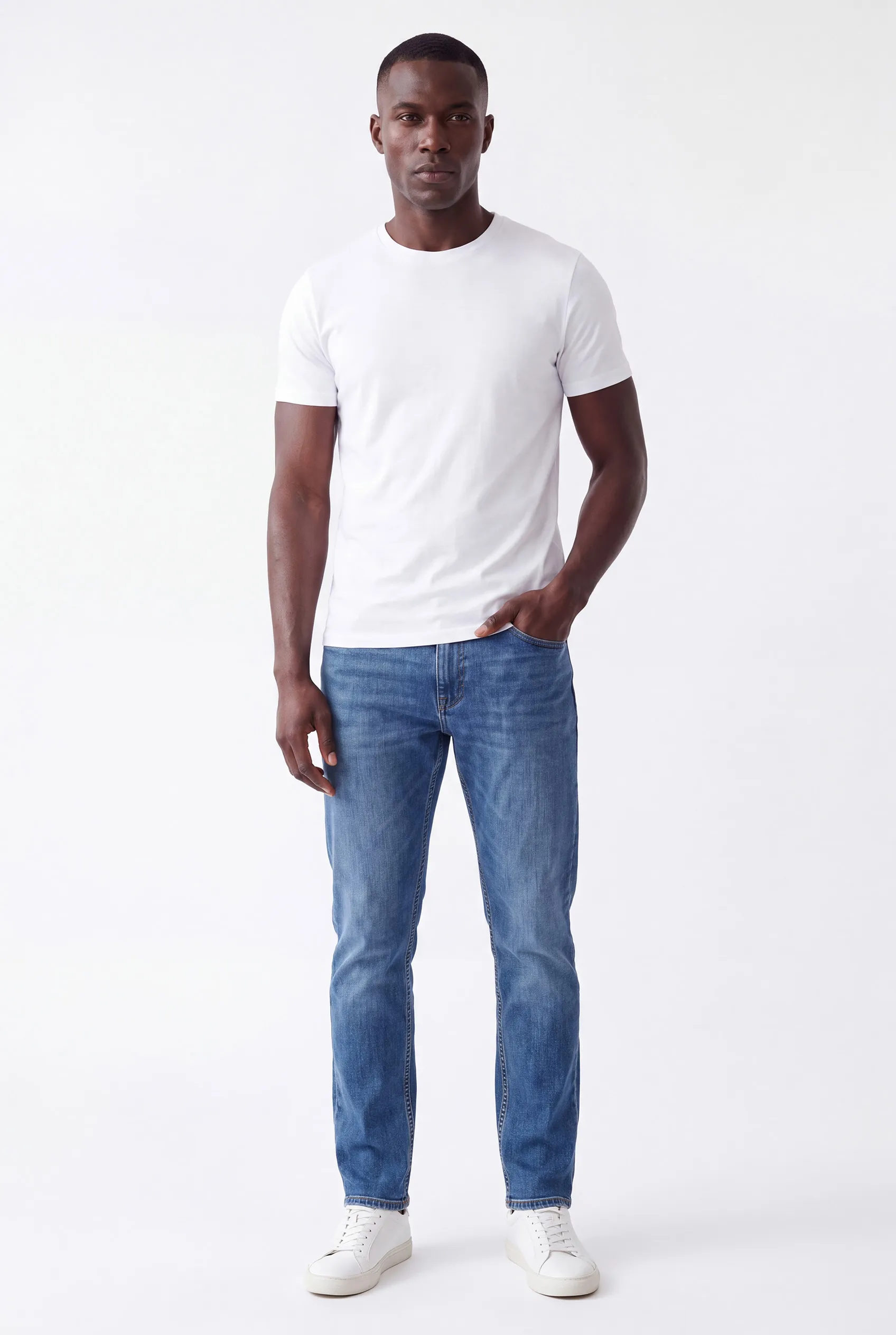 Merkmodel Elian ten voeten uit: man in een wit T-shirt, spijkerbroek en witte sneakers, met één hand in de zak