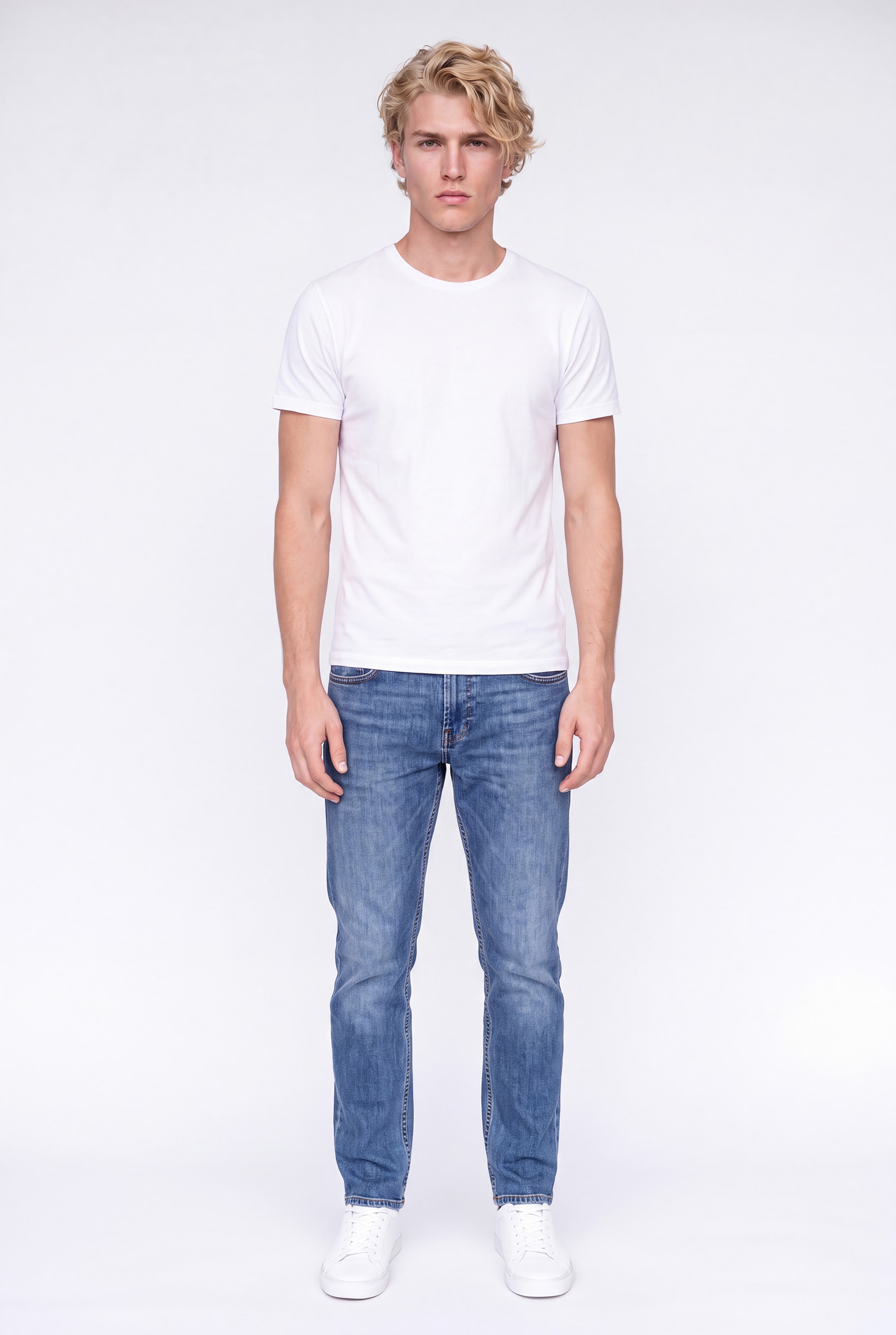 Young man with blond curly hair wearing a plain white t-shirt, blue jeans, and white sneakers, standing against a plain light background facing forward.