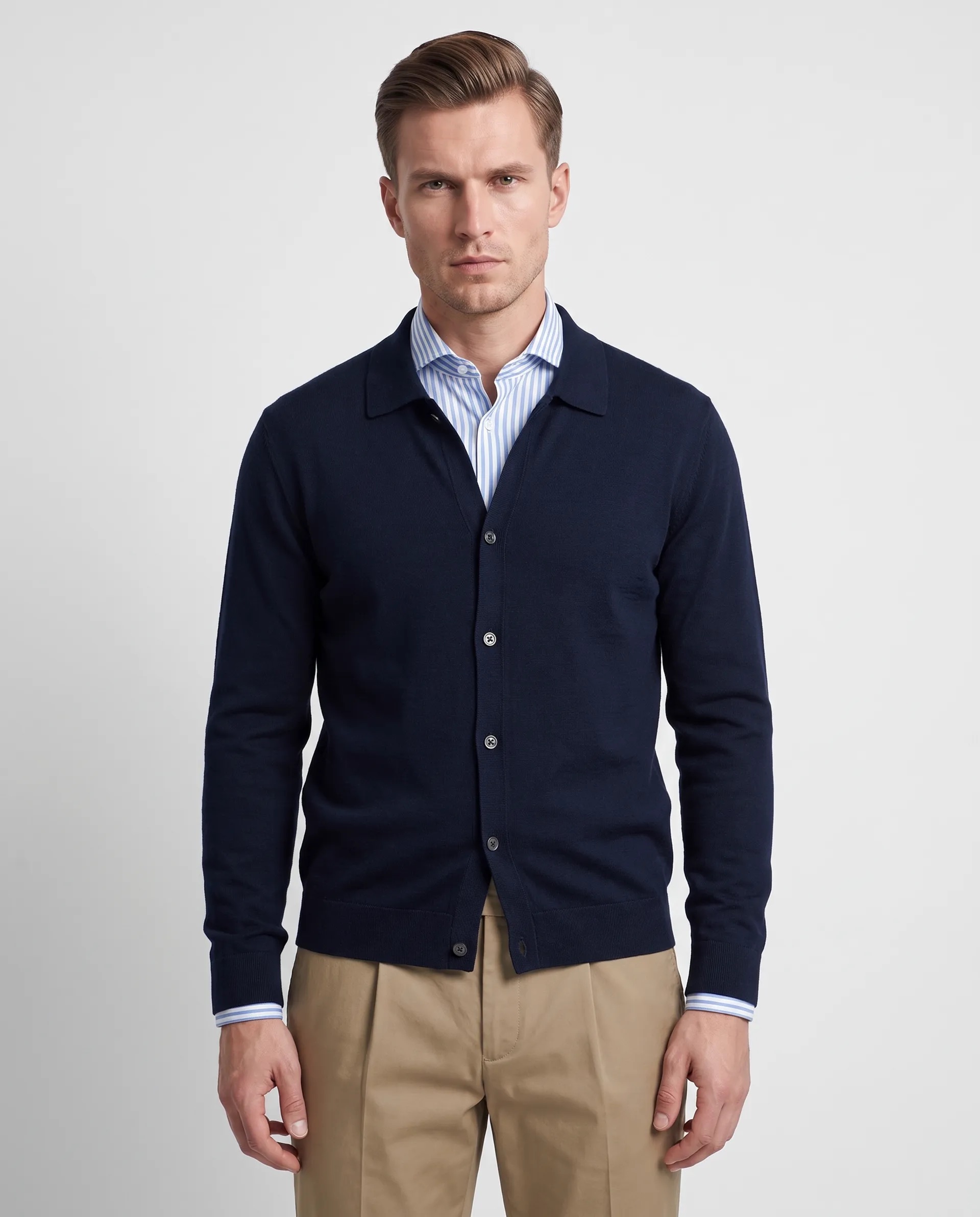 Man wearing a navy blue buttoned cardigan over a blue and white striped dress shirt, paired with beige pants, against a plain light gray background.