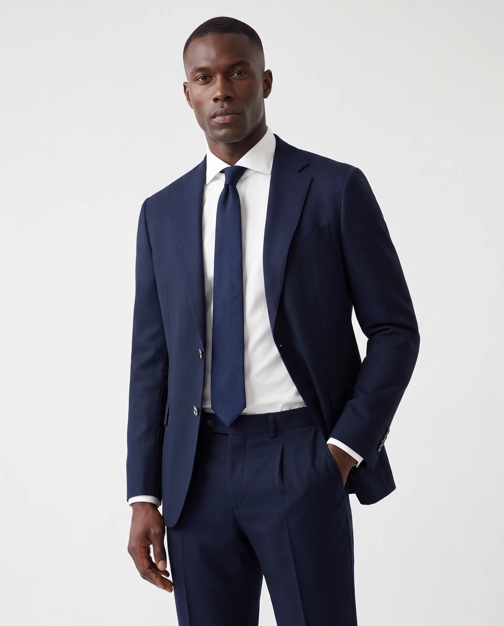 A man wearing a dark navy blue suit with a matching tie and a white dress shirt, standing with one hand in his pocket against a plain light background.