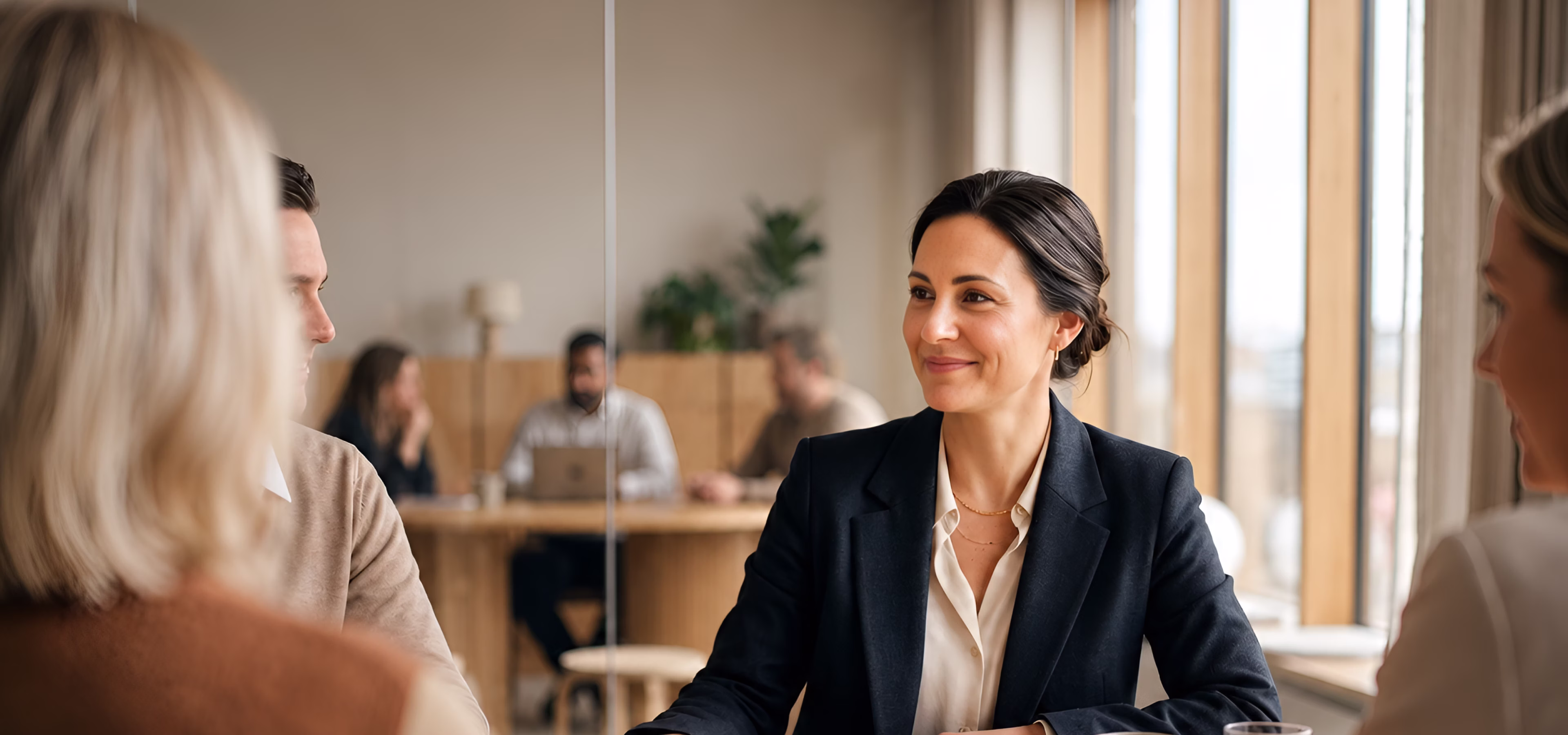 A professional woman in a dark blazer smiles and engages in conversation with colleagues in a modern office setting with large windows and wooden furniture.