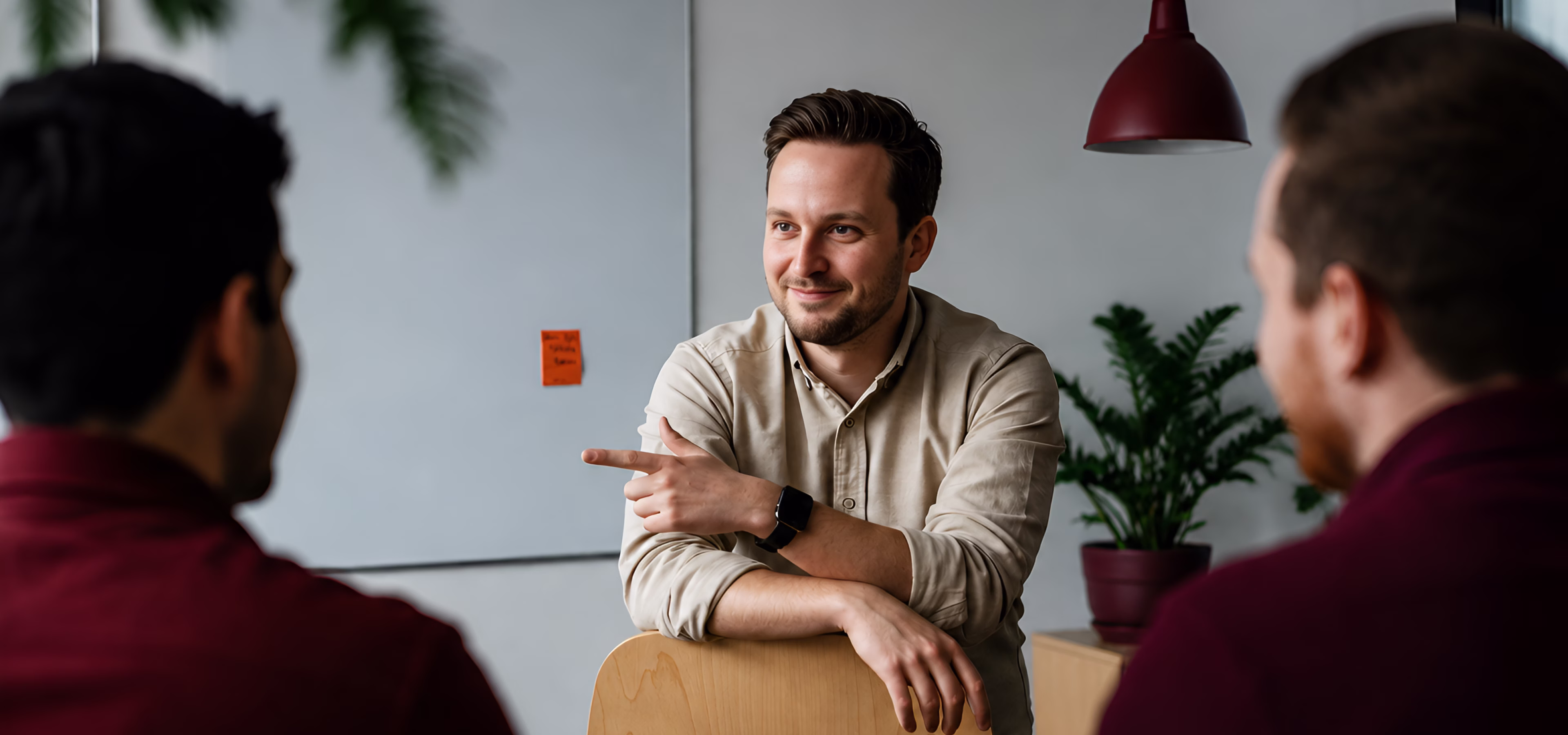 Man in beige shirt smiling and pointing while engaging with two blurred men in a discussion in an office setting with a plant and whiteboard in the background.